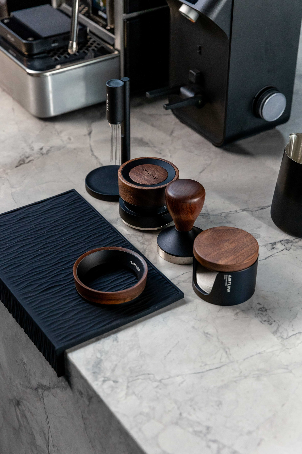 Airflow walnut tamper, dosing ring, WDT tool and tamping mat on a marble bench beside an espresso machine.